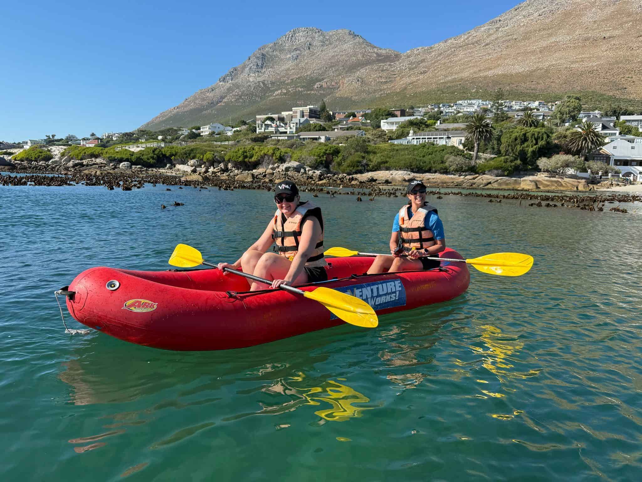 Two people kayaking in a red inflatable kayak on calm waters near Cape Town's coastline, South Africa.
